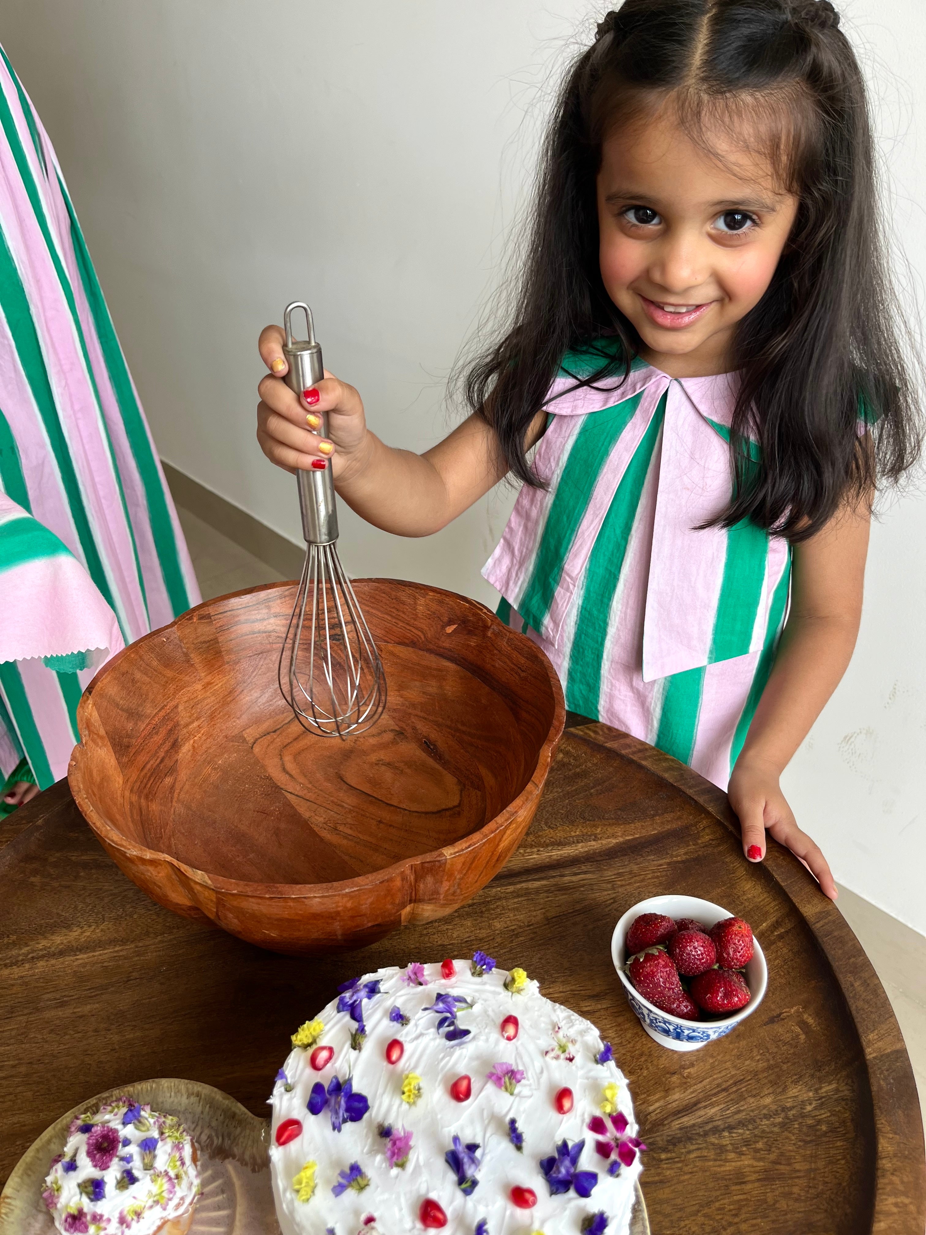 Young girl wearing Imsoo holding a whisk next to a wooden bowl, cake, and strawberries on a table.